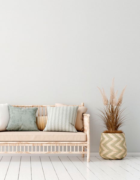 Helles Interieur mit einem Rattansofa, mehreren Kissen und einer Pflanze in einem Korb