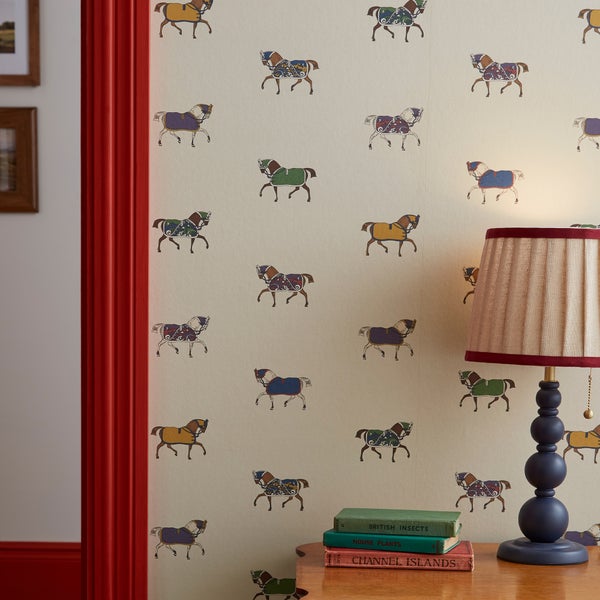 Tapete mit bunten Pferdemotiven an einer Wand, davor ein Holztisch mit Büchern und einer Tischlampe.
