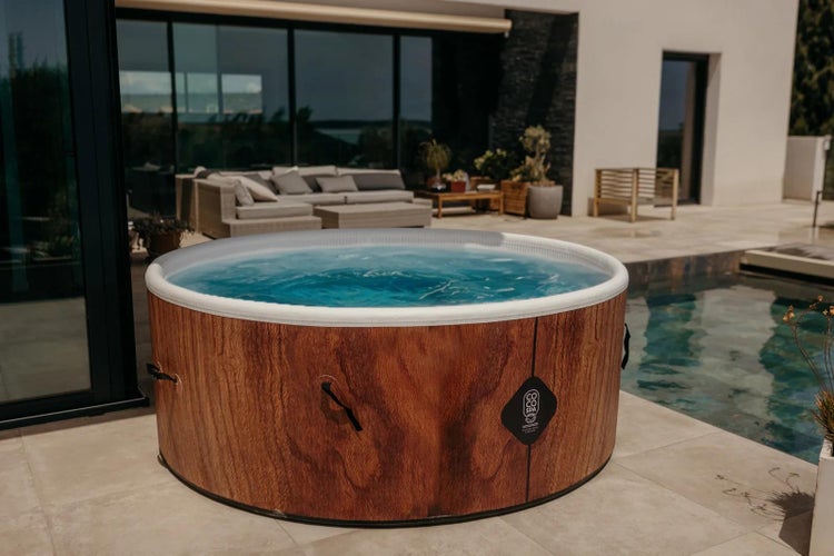 Runder tragbarer Whirlpool in Holzoptik auf einer hellen, geräumigen Terrasse neben einem Pool und modernen Outdoor-Lounge-Möbeln.