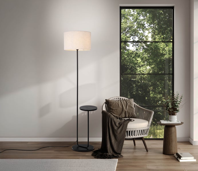 Ein heller Raum mit einer Stehlampe, einem Rattansessel, einem Beistelltisch und einem Fenster mit Blick auf Bäume.