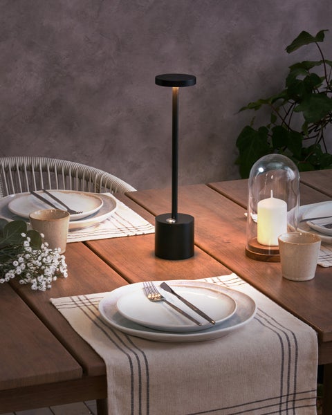 Szene mit einem gedeckten Holztisch mit Geschirr, Kerze und einer schwarzen Tischleuchte.