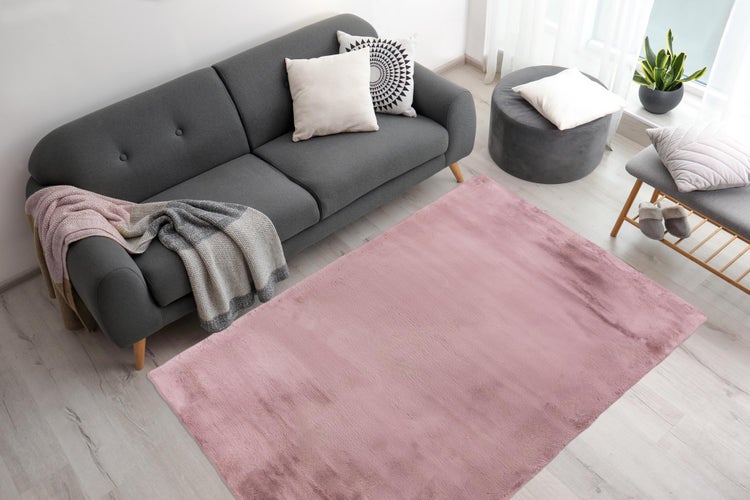 Modernes Wohnzimmer mit einem großen rosa Plüschteppich auf hellem Boden, einem dunkelgrauen Sofa mit Kissen und Wohndecke sowie einem Hocker.