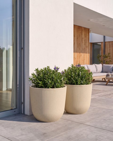 Zwei moderne, geriffelte Pflanzkübel mit Pflanzen auf einer hellen Terrasse mit Bodenfliesen, einer Holzfassade und Gartenmöbeln im Hintergrund.