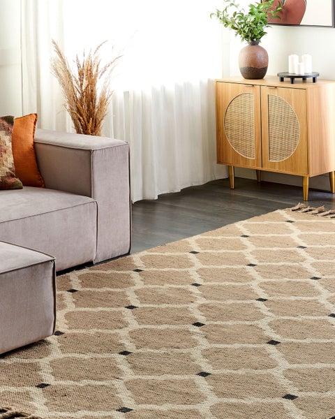 Modernes Wohnzimmer mit gemustertem Teppich in Blau und Grau, Holz-Sideboard, rundem Couchtisch mit Teeservice, Sessel und großen Zimmerpflanzen.