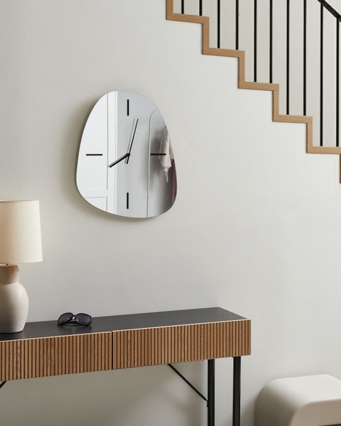 Szene mit einer unregelmäßig geformten Spiegeluhr, einer Kommode mit Lampe und einer Treppe