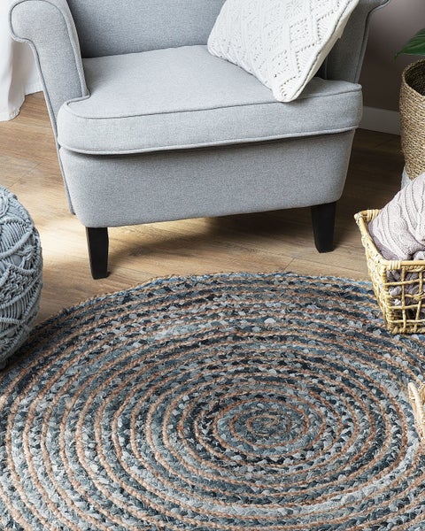 Runder geflochtener Teppich aus Jute und Baumwolle mit konzentrischen Kreisen in einem Wohnzimmer vor einem Sessel auf hellem Holzboden.