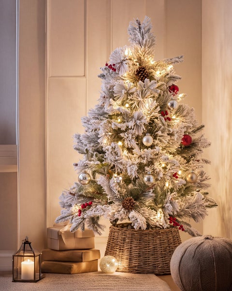 Künstlicher, verschneiter Weihnachtsbaum mit Lichterkette, Kugeln und Beeren in einem Weidenkorb, dekoriert in einem Wohnraum.