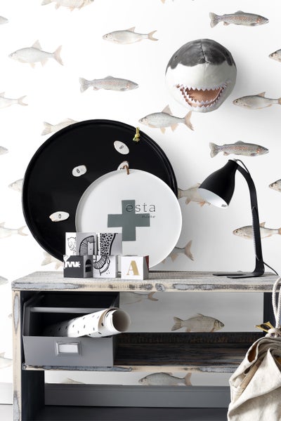 Dekoratives Arrangement mit einer Tischlampe, einem Hai-Wandobjekt und verschiedenen Dekorationselementen auf einem Ablagetisch vor einer Fisch-Tapete.