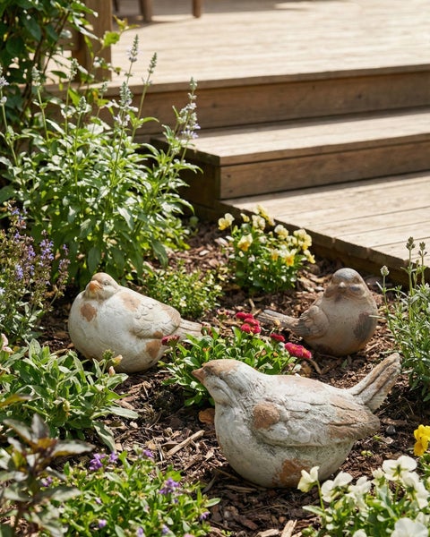 Drei Gartenfiguren in Vogelform in Steinoptik in einem Blumenbeet mit Rindenmulch und verschiedenen Pflanzen vor Holzstufen.