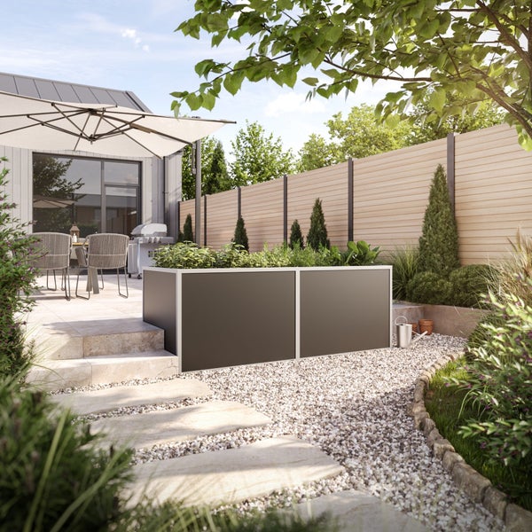Modernes, dunkelgraues Hochbeet aus Metall in einem hellen Garten mit Kiesweg, Steinterrasse und Holzzaun.