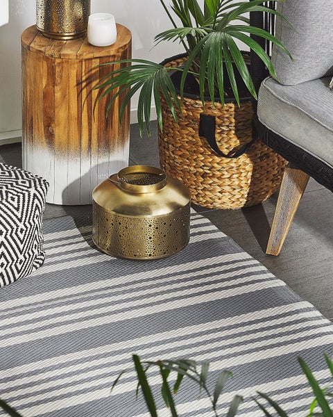 Gemütlicher Außenbereich mit gestreiftem Teppich, goldener Metalllaterne, Beistelltisch aus Holz und Pflanze im Flechtkorb.