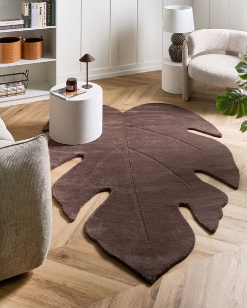 Brauner Teppich in Form eines Monstera-Blatts auf hellem Fischgrätparkett in einem hellen, modernen Wohnzimmer mit Sessel und Regal.