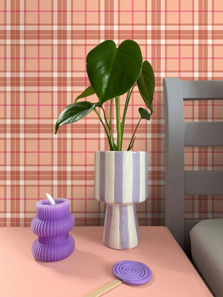 Vliestapete mit Karomuster in einem modernen Schlafzimmer mit einer Monstera-Pflanze im gestreiften Topf, einer gerippten Kerze und einem grauen Bettgestell.