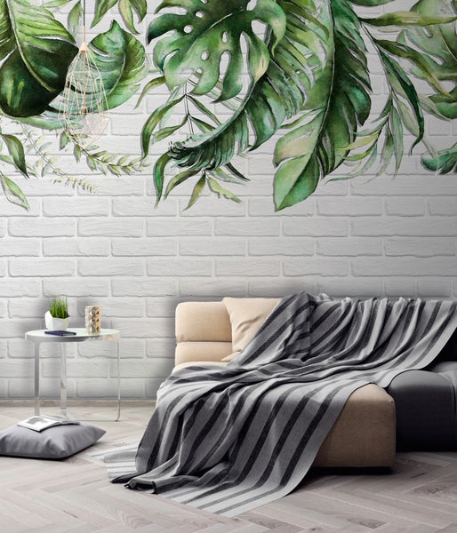 Fototapete mit tropischen Blättern im Aquarellstil an einer weißen Ziegelwand in einem modernen Wohnzimmer mit Sofa und Beistelltisch.