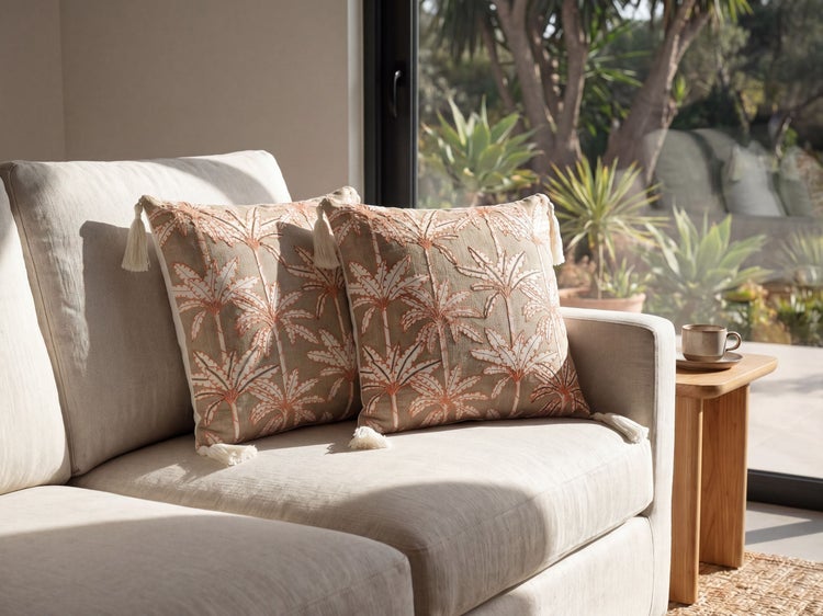 Zwei Zierkissen mit aufgesticktem Palmenmuster und Quasten auf einem Sofa in einem hellen, modernen Wohnzimmer.