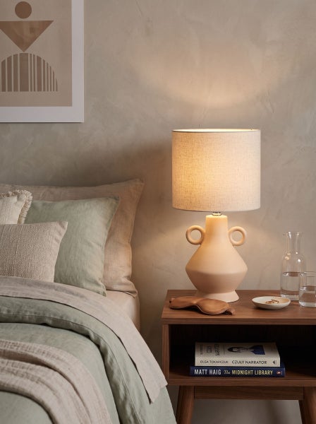 Beleuchtete Tischlampe mit amphorenförmigem Sockel und Lampenschirm aus Stoff auf einem Nachttisch im Schlafzimmer mit Bett und Dekoration.