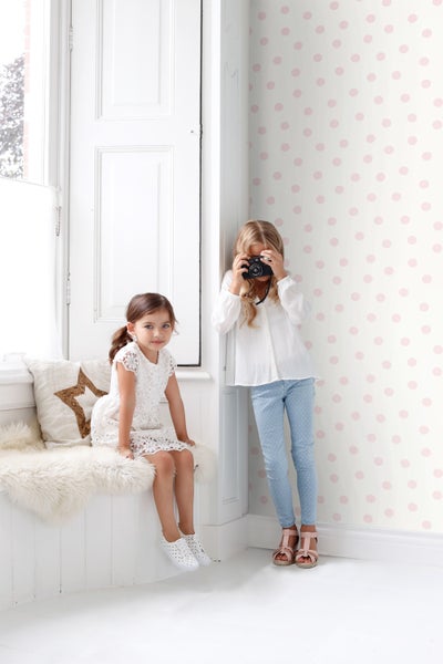 Zwei Mädchen in einem hellen Raum mit gepunkteter Tapete, eines fotografiert mit einer Kamera.