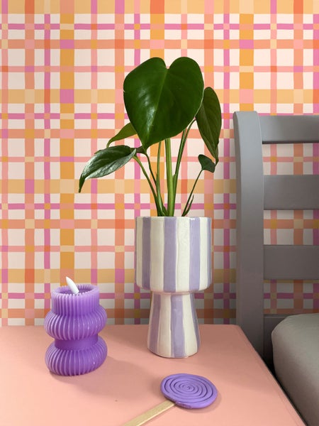 Karierte Vliestapete in Orange und Rosa hinter einem Nachttisch mit Monstera-Pflanze im gestreiften Übertopf und lila Kerze.