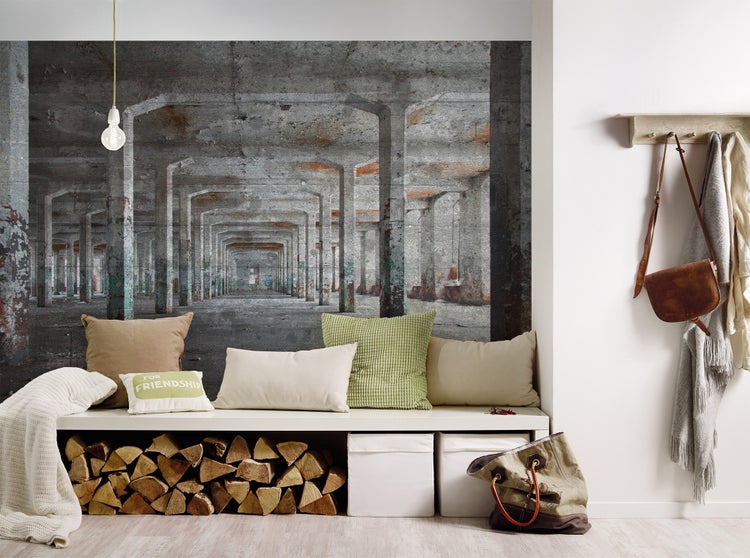 Fototapete mit industrieller Betonhallen-Optik hinter einer weißen Sitzbank mit Kissen, Brennholz und Aufbewahrungsboxen in einem hellen Wohnraum.