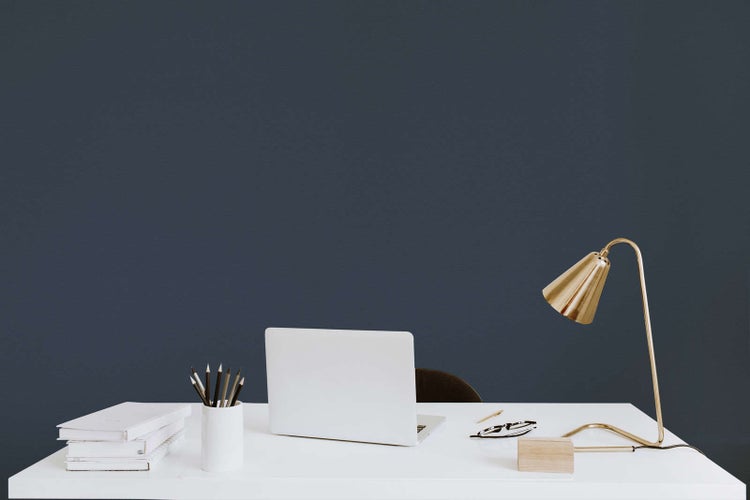 Moderner Arbeitsplatz mit weißem Schreibtisch vor einer dunkelblauen Wand, Laptop, Büchern und einer goldfarbenen Schreibtischlampe.