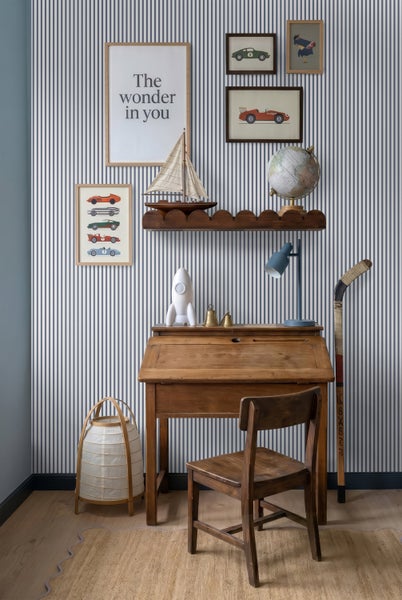 Gestreifte Tapete in einem Kinderzimmer mit Holzschreibtisch, Stuhl und Dekorationsartikeln an einer Wand mit vertikalen Streifen.
