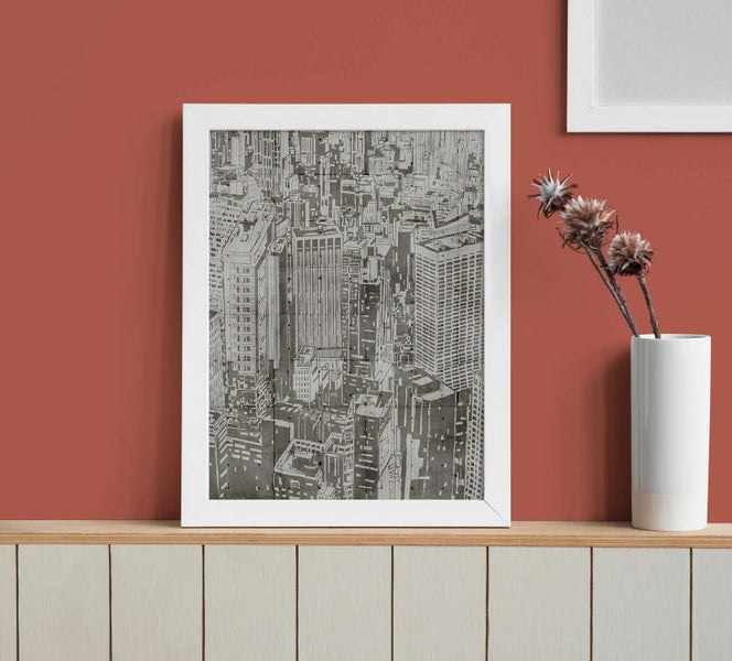 Eingerahmtes Wandbild mit einer schwarz-weißen Stadtansicht von oben in einem weißen Rahmen auf einer Ablage.