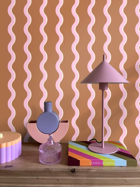 Dekorative Szene mit Lampe, Dose, Vase und Buch auf einem Tisch vor einer gemusterten Wand.