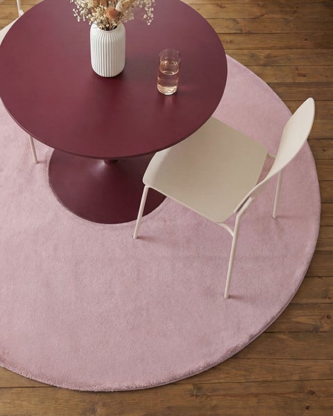 Ein runder, rosafarbener Teppich auf Holzboden, kombiniert mit einem dunkelroten runden Tisch, einem hellen Stuhl und einer Vase.