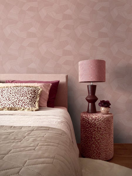 Schlafzimmer mit geometrisch gemusterter Tapete, Bett mit Dekokissen und einer Tischlampe auf einem Pouf.