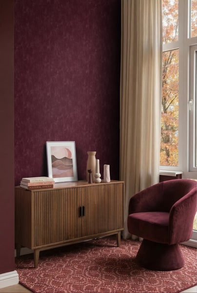 Wohnraum mit weinroter strukturierter Tapete, Sideboard aus Holz, Samtsessel und gemustertem Teppich vor einem Fenster.