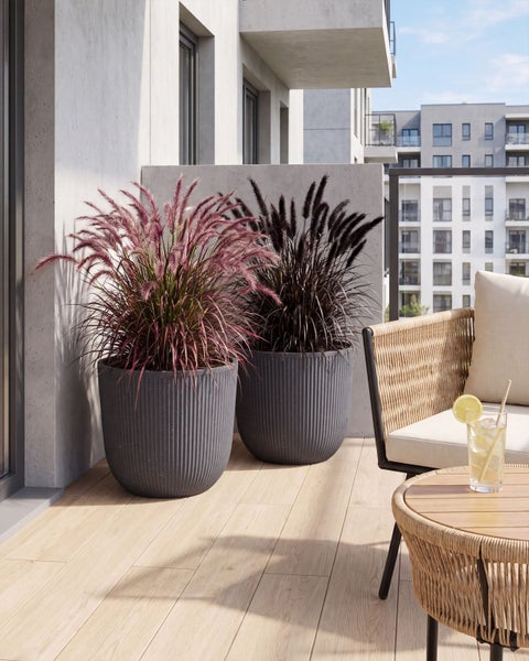 Zwei moderne Pflanzkübel mit Lamellenstruktur und Ziergräsern auf einem Balkon mit Holzoptik Fliesen und Lounge Möbeln.