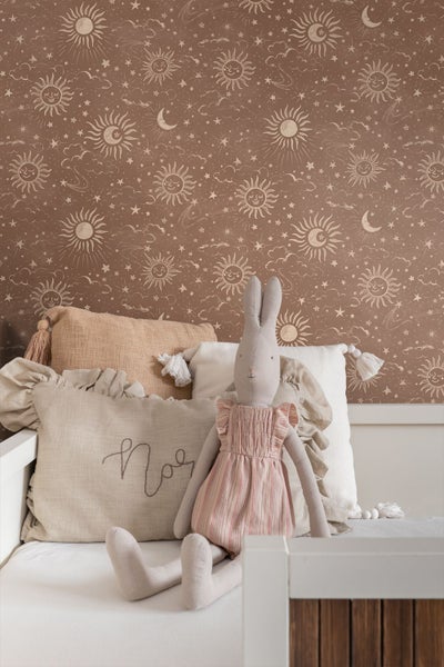 Vliestapete mit Sonnen-, Mond- und Sternenmuster in einem Kinderzimmer mit Kissen und Hasen-Stofftier.