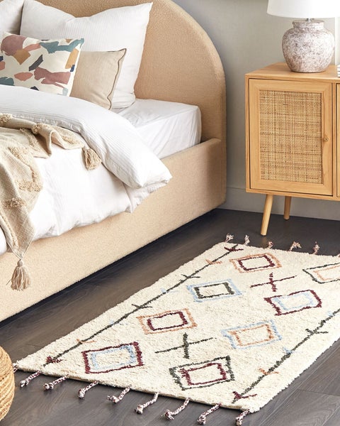 Ein helles, gemütliches Schlafzimmer mit Polsterbett, einem Nachttisch aus Holz mit Rattantür und einem gemusterten Teppich mit Fransen.