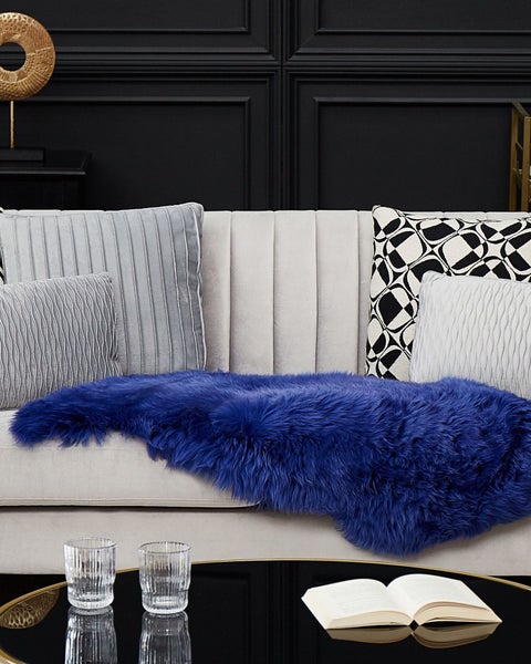Blaues Kunstfell auf einem hellgrauen Sofa mit Zierkissen vor einer dunklen Wand mit Paneelen.