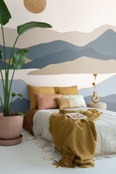 Fototapete mit abstrakten Bergen in einem hellen Schlafzimmer mit Bett, Textilien und einer großen Zimmerpflanze.