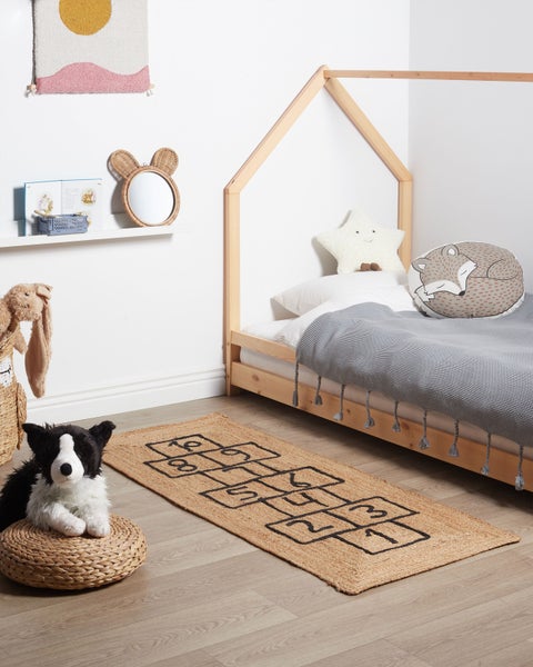 Helles Kinderzimmer mit Hausbett aus Holz, Jute-Teppich mit Hüpfspiel-Muster, Kuscheltieren und dekorativem Spiegel mit Ohren.