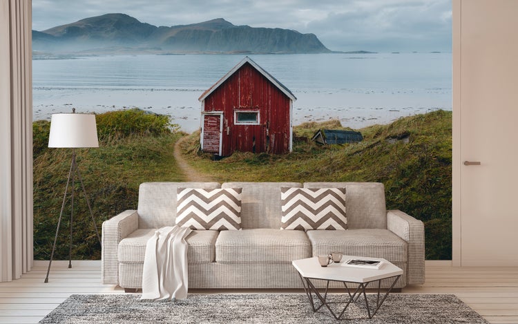 Fototapete mit Küstenlandschaft, roter Hütte und Bergen in einem Wohnzimmer mit Sofa und Stehlampe.
