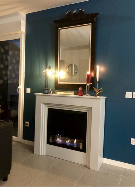 Wohnzimmer mit modernem weißem Kaminumbau und brennendem Ethanol Kamin vor einer Wand in dunklem Blauton mit einem Spiegel darüber.
