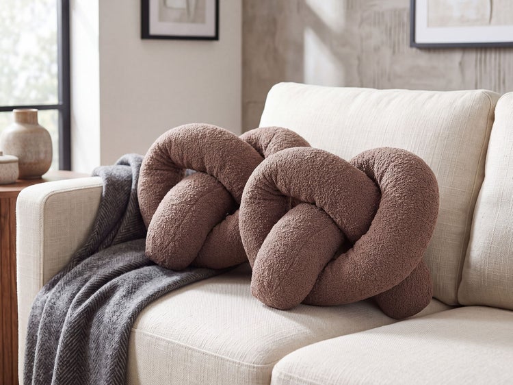 Zwei braune Knoten-Kissen aus Teddy-Stoff auf einem hellen Sofa mit einer grauen Decke in einem modern eingerichteten Wohnzimmer.