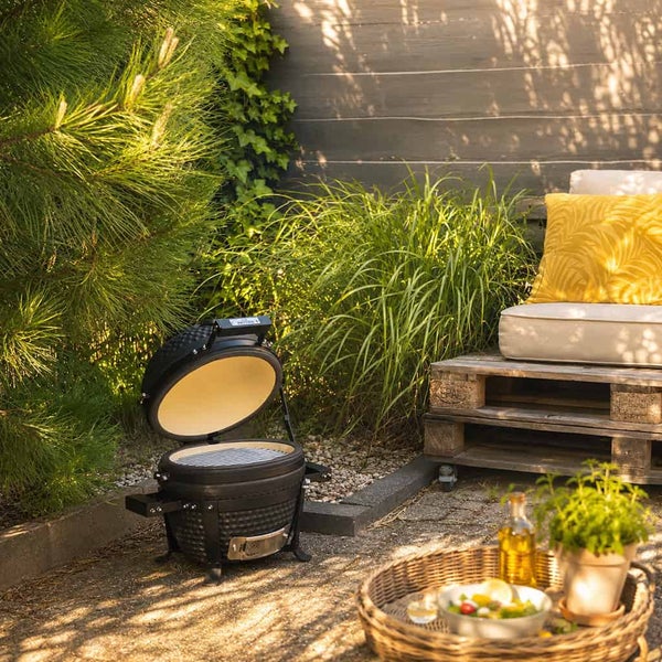 Kompakter Kamado-Grill aus Keramik in einem hellen Garten neben Palettenmöbeln und grünen Pflanzen.