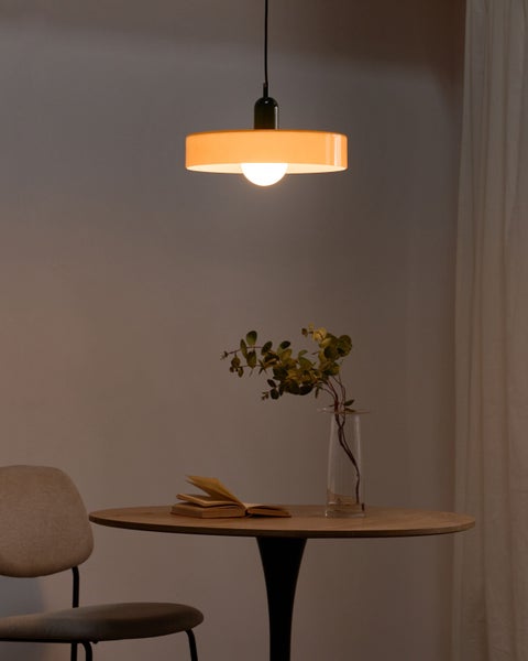 Moderne Pendelleuchte mit orangefarbenem Schirm über einem runden Holztisch mit Buch und Vase in einem schlichten, hellen Raum.