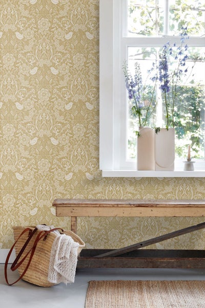 Vliestapete mit floralem Muster und Vögeln an einer Wand hinter einer rustikalen Holzbank und einem Fenster.