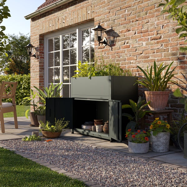 Dunkler Gartenschrank aus Metall mit integriertem Hochbeet vor einer Backsteinwand, umgeben von Pflanzen auf einer Terrasse.