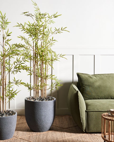 Dekoratives Interieur mit Kunstbambus in strukturierten Töpfen neben einem grünen Sofa auf einem Teppich.