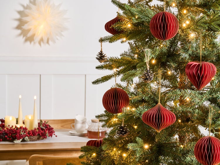 Weihnachtsbaum dekoriert mit roten Papierornamenten in Wabenoptik und Kiefernzapfen vor einem festlich gedeckten Tisch mit Kerzen.