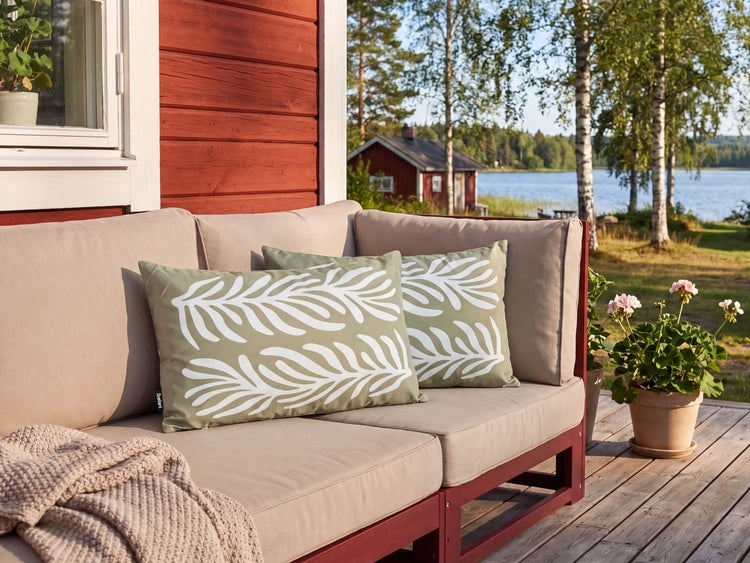 Gartensofa mit Kissen mit Blattmuster auf einer Holzterrasse vor einem Seehaus.