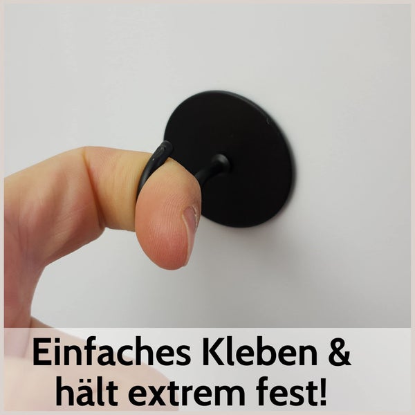 Selbstklebender Wandhaken in Schwarz mit runder Basis, der unter Belastung durch einen Finger die starke Haftkraft zeigt.