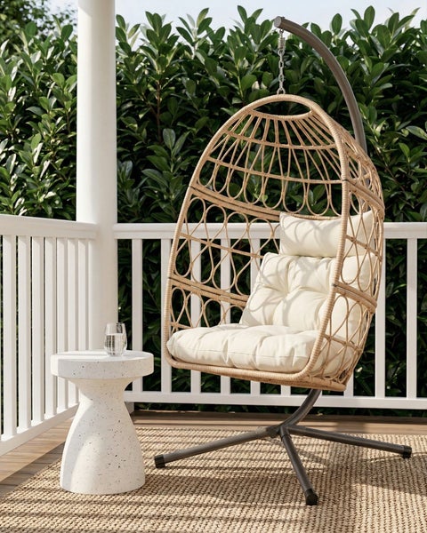 Hängesessel in Eiform aus Polyrattan mit hellen Kissen auf einer Terrasse neben einem weißen Beistelltisch.
