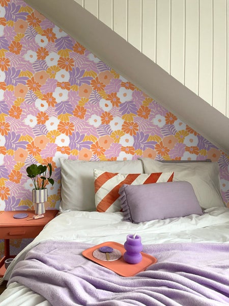 Tapete mit floralem Retro-Muster in Lila und Orange an einer Wand in einem Schlafzimmer mit Bett und Dekoration.