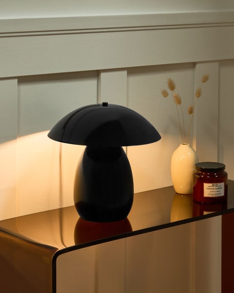 Moderne schwarze Tischleuchte in Pilzform auf einem gläsernen Sideboard neben einer Vase und einer Kerze in einem hellen, organisierten Raum.